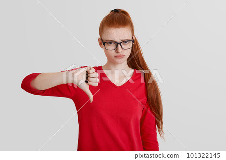 Portrait of pretty young female shows dissaproval and dissagreement, keeps thumb down, has opposite opinion, being in bad mood, expresses negative emotions, isolated over white background Portrait of pretty young female shows dissaproval and dissagreement, keeps thumb down, has opposite opinion, being in bad mood, expresses negative emotions, isolated over white background 101322145