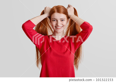 Positive redhead freckled female has long hair, makes pony tails, being pleased to pose at camera, wears red casual sweater, isolated over white background. People, beauty and youth concept. 101322153