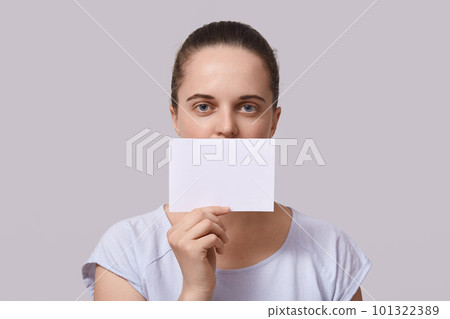 Closeup portrait of serious young female wearing white casual t shirt, covering her mouth with empty card with copy space for inscription, advertisement posing against studio wall. Advertising concept 101322389