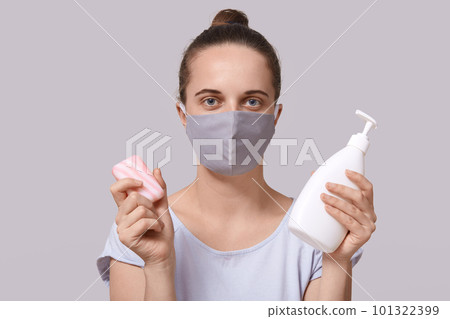 Studio shot of confident serious cute young female holding disinfectant alcohol gel and soap, sticking to rules, protecting herself from coronavirus, wearing antibacterial mask. Covid19 concept. 101322399