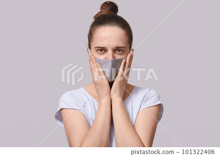 Indoor shot of woman wearing casual clothing and protective mask, keeping palms on cheek, posing isolated over white background, female being afraid of being infected corona virus. Health care concept 101322406