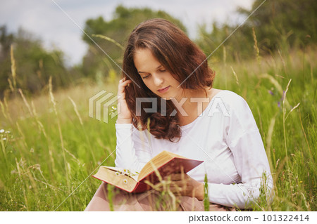Portrait of lovely beautiful female with attractive look reads book in open air, sits in green field, enjoys summer sunny weather and wonderful nature view. Woman likes calm atmosphere while reading Portrait of lovely beautiful female with attractive look reads book in open air, sits in green field, enjoys summer sunny weather and wonderful nature view. Woman likes calm atmosphere while reading 101322414