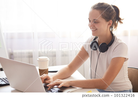 Side view of young woman sitting at table and enjoying coffee while working. Female keyboarding on laptop, lady wearing white casual t shirt with headphones around neck, using wireless internet. 101322416