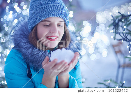 Outdoor shot of glad smiling beautiful woman has red lips and attractive appearance, holds white snow in hands, admires winter wonderful magic night city and frosty weather, plays with snow. Outdoor shot of glad smiling beautiful woman has red lips and attractive appearance, holds white snow in hands, admires winter wonderful magic night city and frosty weather, plays with snow. 101322422