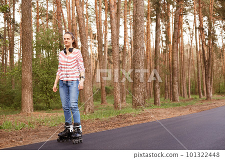 Attractive slim woman wearing jeans and striped shirt,having headphones around neck, rollerblading in forest, breathing fresh air, being in good, expressing positive emotions. Sport concept. Attractive slim woman wearing jeans and striped shirt,having headphones around neck, rollerblading in forest, breathing fresh air, being in good, expressing positive emotions. Sport concept. 101322448