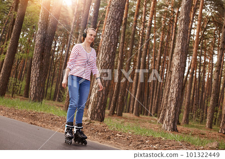 Outdoor photo of attractive magnetic woman having positive facial expression, having roller skates on legs, listening to music, wearing headphones, jeans and striped sweatshirt. Hobby concept. 101322449