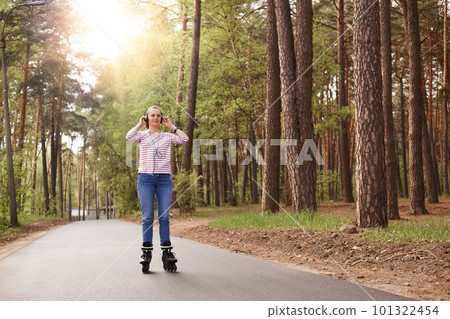 Attractive sporty woman wearing casual attire rollerblading and listening to music, keeping hands on her headphones, spending time with her family in forest. Winsome girl rinking in open air. Attractive sporty woman wearing casual attire rollerblading and listening to music, keeping hands on her headphones, spending time with her family in forest. Winsome girl rinking in open air. 101322454
