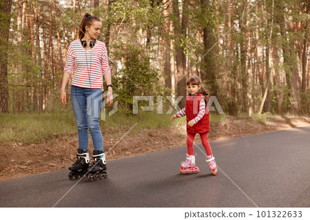 Outdoor shot of young mummy with her cute daughter rollerblading in forest on asphalt road, mom looking at her child, wearing casual outfits, family spending time together in open air. People concept Outdoor shot of young mummy with her cute daughter rollerblading in forest on asphalt road, mom looking at her child, wearing casual outfits, family spending time together in open air. People concept 101322633