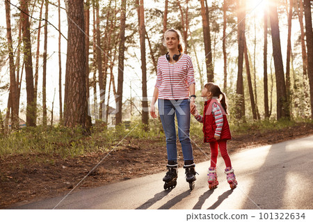 Happy woman enjoying to spend time with her charming little daughter, wearing casual attires, posing in forest, riding rolling skates on asphalt road in park. 101322634