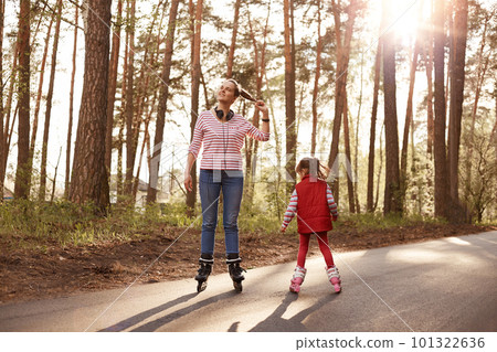 Outside picture of cheerful energetic young female rollerskating with her little daughter, enjoying nature, being at forest, having headphones around neck, leading active lifestyle. Hobby concept. Outside picture of cheerful energetic young female rollerskating with her little daughter, enjoying nature, being at forest, having headphones around neck, leading active lifestyle. Hobby concept. 101322636
