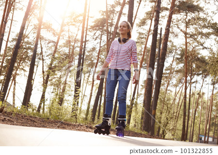 Outdoor photo of attractive positive woman having headphones around neck, enjoying spring sunny days, being around nature, having peaceful facial expression, being in good mood. Hobby concept. 101322855