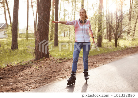 Happy young girl wearing casual striped shirt and jeans, roller skating in park, enjoying beautiful nature and fresh air, spreading hand in front of her. 101322880