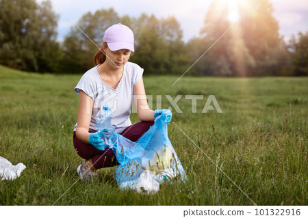 Attractive lady wearing white casual t shirt, baseball cap and trousers, picking up used plastic bag, cleaning dirty meadow, female looks concentrated, helping planet. 101322916