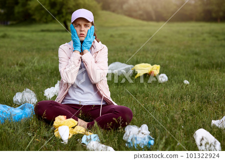 Astonished and shocked woman sitting in park on green grass with palms on cheeks, wearing casually, surrounded with waste, needs to pick up all garbage. Astonished and shocked woman sitting in park on green grass with palms on cheeks, wearing casually, surrounded with waste, needs to pick up all garbage. 101322924