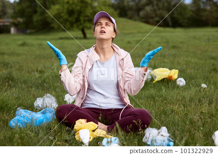 Young attractive lady being in despair sitting on green lawn with spreading hands and looking at sky, being tired and shocked of nature pollution, picking up trash in meadow. 101322929