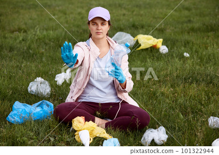 Activist picking up trash in field, sitting on green grass and holding plastic bottle, showing stop sign with her palm, female calls for saving our planet from pollution,solving ecological problems. Activist picking up trash in field, sitting on green grass and holding plastic bottle, showing stop sign with her palm, female calls for saving our planet from pollution,solving ecological problems. 101322944