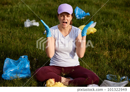 Lady in panic sitting on ground in field, screaming, being shocked of pollution, needs to pick up lots of trash, wearing casual attire, having angry facial expression. 101322949