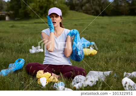 Tired astonished female picking up litter on green meadow, sitting on ground with opened mouth and keeping palm in latex gloves on cheek, being shocked from pollution and human irresponsibility. Tired astonished female picking up litter on green meadow, sitting on ground with opened mouth and keeping palm in latex gloves on cheek, being shocked from pollution and human irresponsibility. 101322951