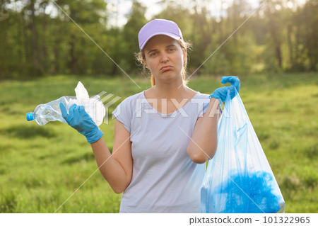 Outdoor portrait of woman volunteer picking up garbage in meadow or park for clean planet, environmentally and ecology friendly, looks at camera with pout lips, holding plastic bottle and garbage bag. Outdoor portrait of woman volunteer picking up garbage in meadow or park for clean planet, environmentally and ecology friendly, looks at camera with pout lips, holding plastic bottle and garbage bag. 101322965