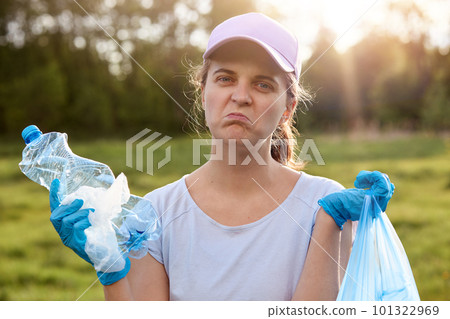 Lady with twisted face wearing blue latex gloves, holding rubbish in hands, looks at camera with upset facial expression, wants to clean up planet from litter and reusing waste, ecological problems. 101322969