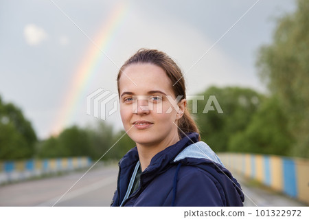 Outdoor portrait of young woman wearing dark blue jacket posing in open air and looking at camera with dreamily expression, standing with rainbow on background. 101322972