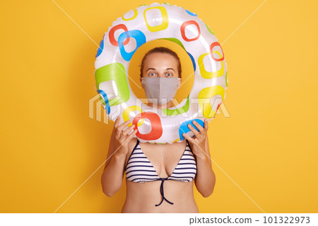 Woman in striped swimsuit looking through rubber ring, wearing hygienic face mask to prevent virus on resort beach, rest and summer vacation with means of protection her health. 101322973