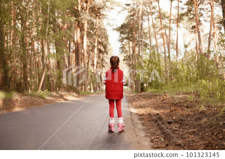 Picture of active energetic little girl wearing red suit, rollerskating during holidays, enjoying nature, breathing fresh air, being alone on road, spending time in forest, standing back to camera. 101323145