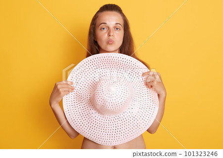 Young beautiful woman with dark hair without clothes, hides behind big straw hat isolated over yellow background, keeping lips rounded like showing kiss gesture. 101323246