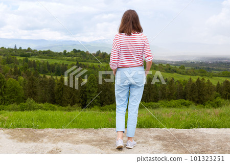 Woman standing in mountain, panoramic view of mountain range, trees. green grass. blue sky, female wearing casual trousers and white t shirt with red stripes enjoing beautiful nature. People concept. 101323251