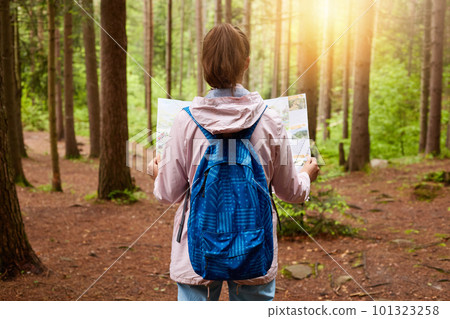 Back view of beautiful young girl with map in hands standing in forest. Tourist wearing rose jacket and blue bag trying to find her way, taking part in quest. Concept of finding way and traveling. Back view of beautiful young girl with map in hands standing in forest. Tourist wearing rose jacket and blue bag trying to find her way, taking part in quest. Concept of finding way and traveling. 101323258
