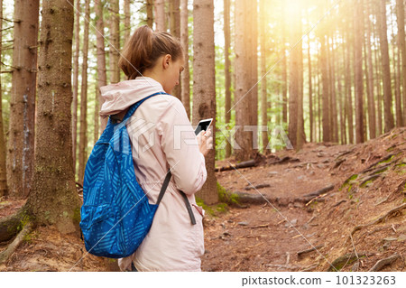 Image of woman backpacker trying to find her way, using mobile phone in forest, female going for walk in spring season, attractive lady dressed casually. Traveling and having adventures concept. 101323263