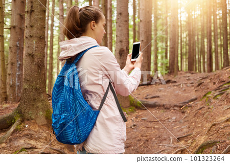Image of active experienced tourist wearing pink jacket and blue backpack, holding her smartphone, trying to find connection, taking photos of forest, being around nature. Travelling concept. 101323264