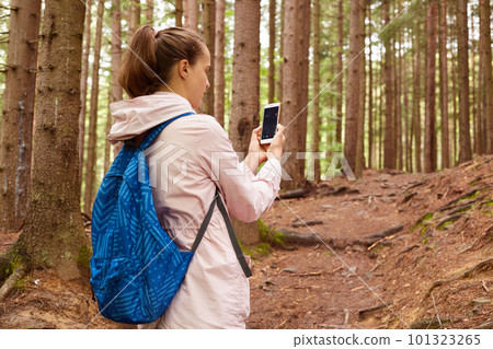 Profile of hiker woman wandering in forest, walking up slope, holding smartphone in both hands, switching it on, looking for connection, wearing pink jacket and blue backpacker. Travelling concept. 101323265