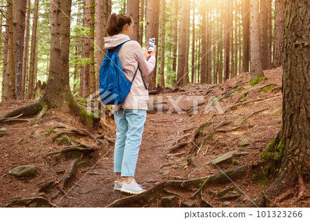 Image of experienced travel blogger standing in middle of forest around huge trees, holding her smartphone in both hands, taking photos for social networking sites, going on trip alone. Traveling Image of experienced travel blogger standing in middle of forest around huge trees, holding her smartphone in both hands, taking photos for social networking sites, going on trip alone. Traveling 101323266