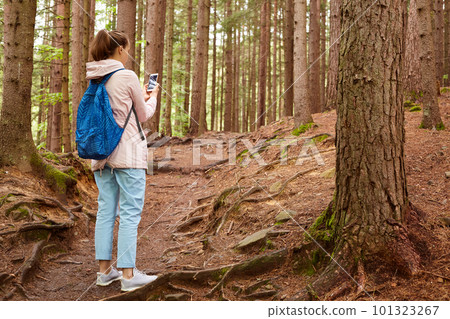 Back view of experienced traveller standing in middle of forest, getting lost, looking for way out, using her smartphone, taking photo, recording video for travel blog, spending time with nature. Back view of experienced traveller standing in middle of forest, getting lost, looking for way out, using her smartphone, taking photo, recording video for travel blog, spending time with nature. 101323267