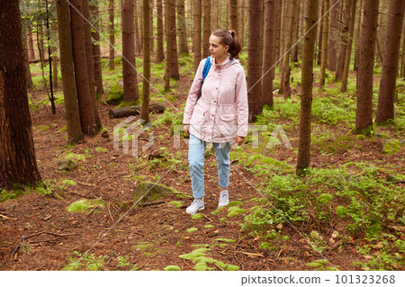 Outdoor shot of enthusiastic tourist having camping tour, searching for right path, looks delighted, wearing jeans, pink jacket and blue backpacker, having pony tail. Leisure activities concept. 101323268