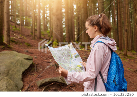 Attractive energetic young lady holding map in both hands, looking attentively at map, getting lost in forest, improving orientating skills, fond of active rest, being concentrated. Active lifestyle. 101323280