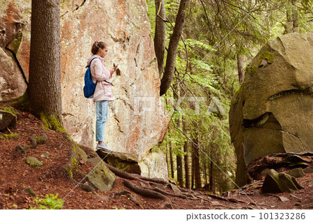 Profile of young attractive tall traveller holding mobile phone in hands, looking attentively at screen, trying to find connection, having orientating problems, taking photos, walking between stones. 101323286