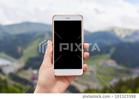 Outdoor image of one hand holding and showing white smartphone with blank black desktop screen with blur green mountains on background, mobile phone switched off. Copyspace for advertisement. 101323398