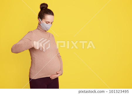 Young pregnant girl in a protective mask points her hands at her stomach against yellow background, expectant mother with hair bun, looking at her stomach, dresses casually. Young pregnant girl in a protective mask points her hands at her stomach against yellow background, expectant mother with hair bun, looking at her stomach, dresses casually. 101323495