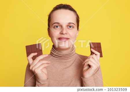 Pregnant woman holding two bars of chocolate in both hands and looking directly at camera with satisfied facial expression, posing isolated over yellow background, wants to eat junk food. Pregnant woman holding two bars of chocolate in both hands and looking directly at camera with satisfied facial expression, posing isolated over yellow background, wants to eat junk food. 101323509