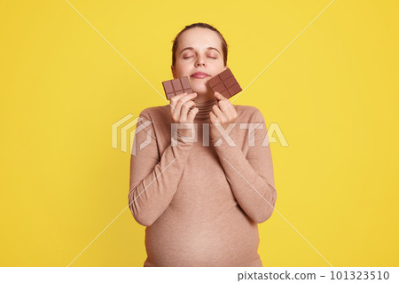 Satisfied expectant mother standing against yellow wall with closed eyes, holding bars of chocolate in both hands, enjoying eating eat sweets, wearing beige sweater. Satisfied expectant mother standing against yellow wall with closed eyes, holding bars of chocolate in both hands, enjoying eating eat sweets, wearing beige sweater. 101323510