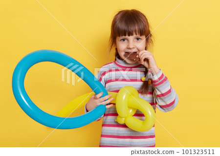 Portrait of happy little girl eating chocolate and holding balloon figure in hands, looking at camera with satisfied expression, wearing casual striped turtleneck, isolated over yellow background. Portrait of happy little girl eating chocolate and holding balloon figure in hands, looking at camera with satisfied expression, wearing casual striped turtleneck, isolated over yellow background. 101323511