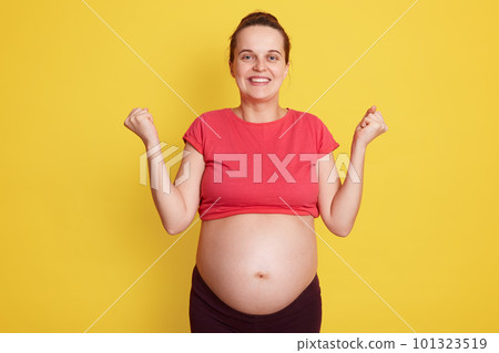 Pregnancy, motherhood, expectation baby. Close up portrait of happy pregnant woman with big bare belly standing with excitement expression isolated over yellow background, keeping fists clenched. 101323519