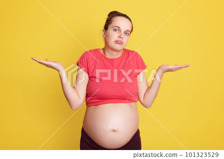 Young beautiful female expecting baby, stands isolated over yellow background, being clueless and confused with open arms, having no idea, shrugging shoulders, shows helpless gesture. Young beautiful female expecting baby, stands isolated over yellow background, being clueless and confused with open arms, having no idea, shrugging shoulders, shows helpless gesture. 101323529