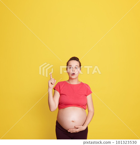 Surprised pregnant woman pointing finger up isolated over yellow background, lady with widely opened mouth touching her bare tummy, expectant mother. Copy space for add. 101323530