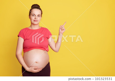 Caucasian young pretty pregnant woman touching belly and pointing aside isolated over yellow background, expectant mother with bare tummy wearing red t shirt. Copy space for advertisement. Caucasian young pretty pregnant woman touching belly and pointing aside isolated over yellow background, expectant mother with bare tummy wearing red t shirt. Copy space for advertisement. 101323531