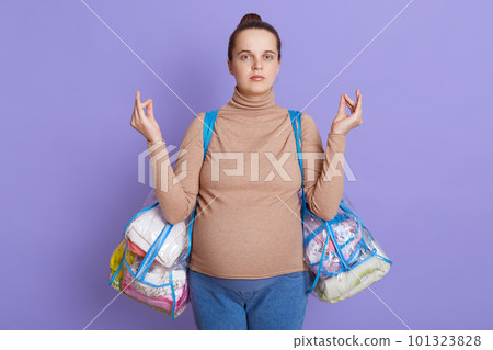 Beautiful young pregnant woman with hair bun meditates before going to maternity home to give birth, trying to relax, holding stuff for baby in two bags, posing isolated over lilac background. Beautiful young pregnant woman with hair bun meditates before going to maternity home to give birth, trying to relax, holding stuff for baby in two bags, posing isolated over lilac background. 101323828