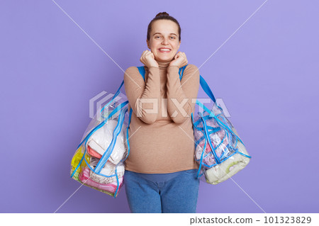 Young beautiful pregnant woman standing on lilac background with happy facial expression, keeping fists under chin, looking at camera with toothy smile, wearing casually, holding bags. Young beautiful pregnant woman standing on lilac background with happy facial expression, keeping fists under chin, looking at camera with toothy smile, wearing casually, holding bags. 101323829