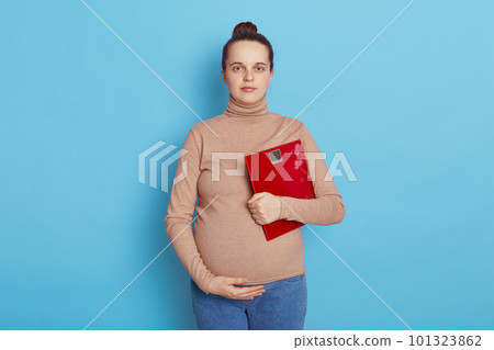 Pretty young pregnant woman holding floor scales in hands, future mother with hair bun touching belly and looking directly at camera, expectant mommy against blue wall. 101323862